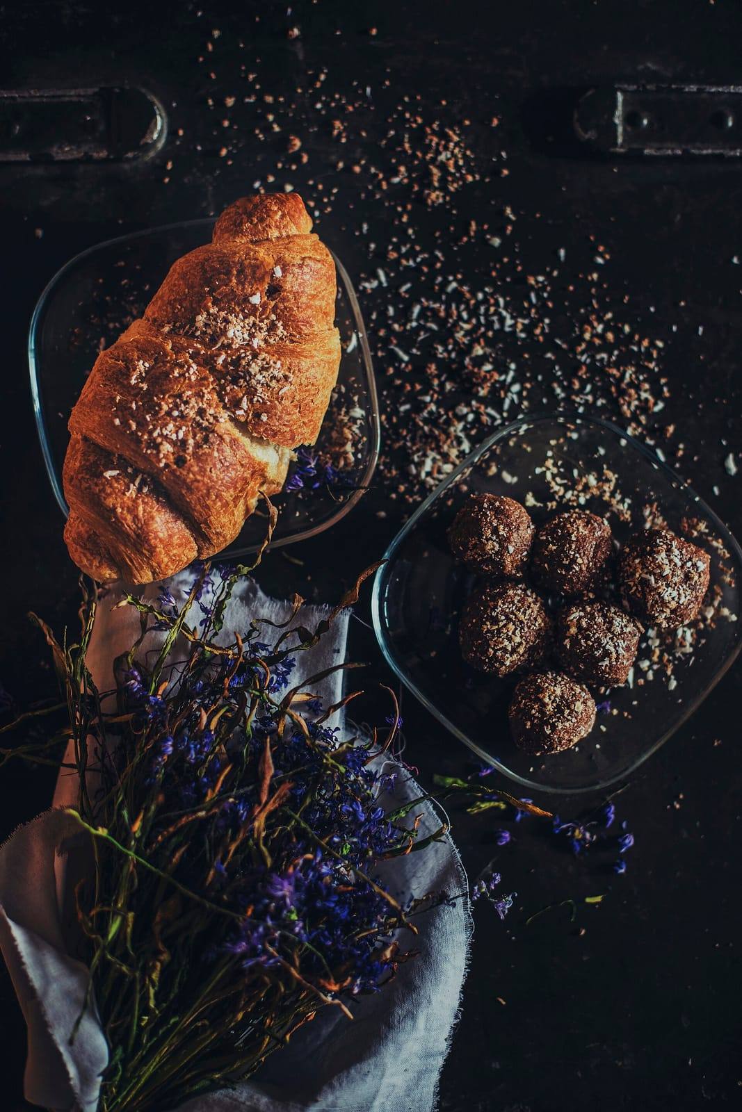 A single golden croissant on a dark wooden table beside a bowl of chocolate truffles
