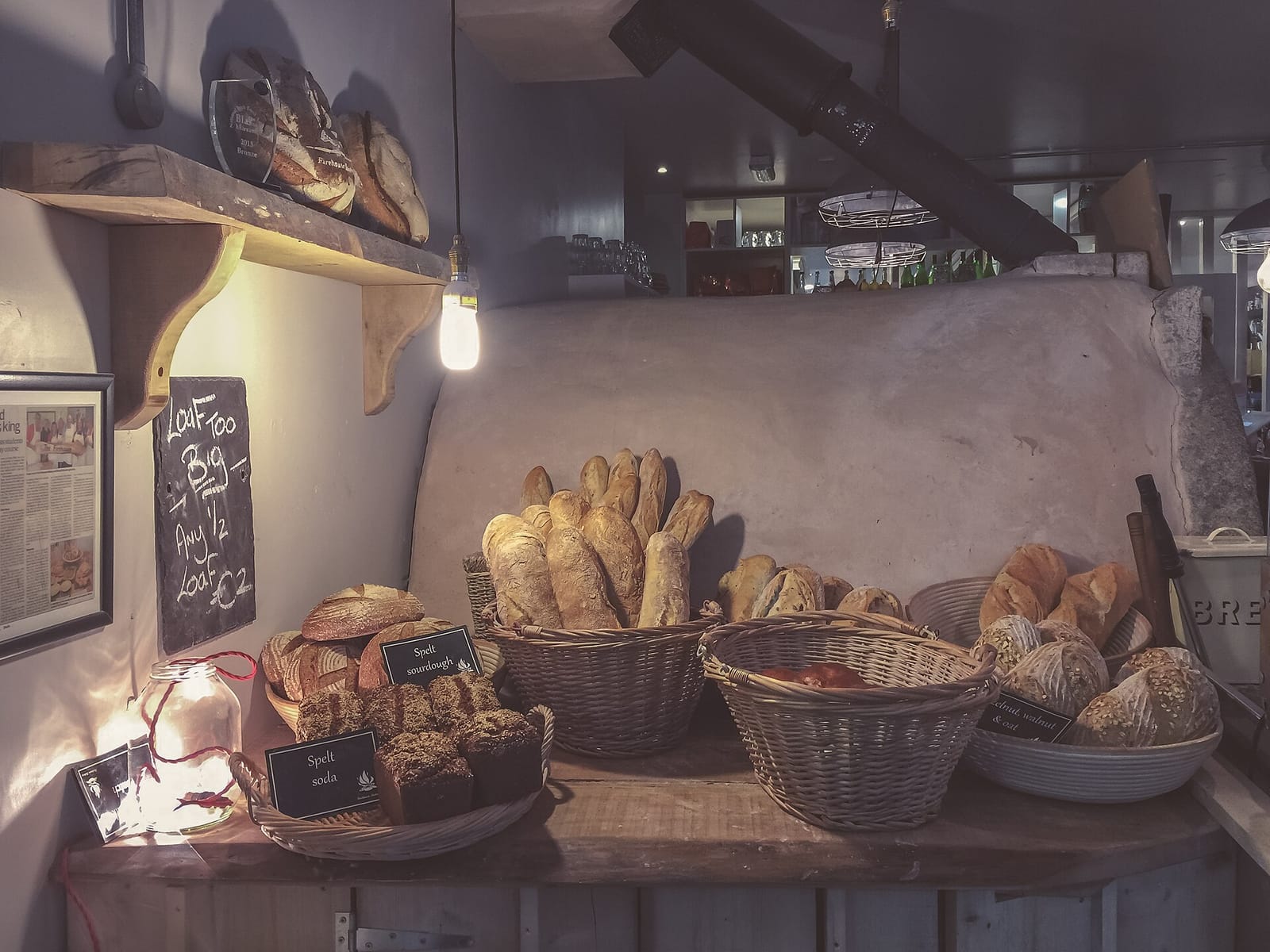 Round and long loaves of crusty bread stacked in wicker baskets on a wooden bakery shelf under a warm lamp