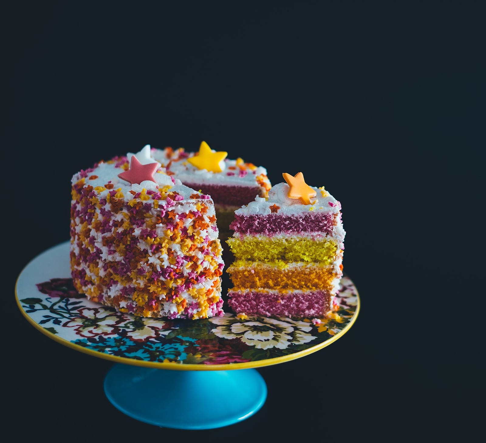 A sprinkle-covered layer cake with star candies on top, one slice cut out to show pink, yellow and orange sponge
